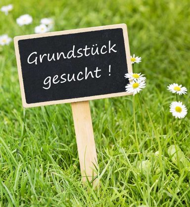 Ein Holzschild mit der Aufschrift "Grundstück gesucht!" auf grünem Rasen.
