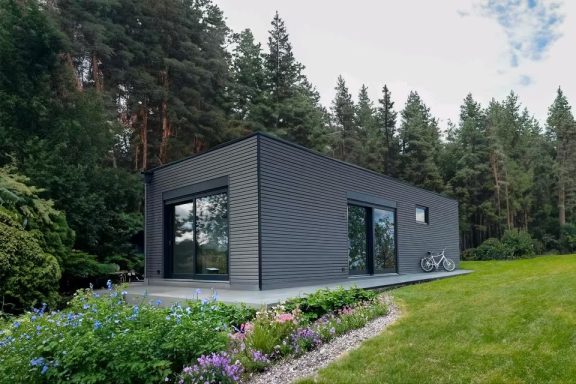 Moderne, dunkelgraue Fertighaus umgeben von Bäumen und bunten Blumen.